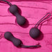 The Je Joue Ami Kegel Ball Exerciser Set, featuring three black silicone kegel balls with string loops, is arranged on a textured pink surface. Two balls are elongated with curves, and one is spherical, gracefully contrasting with the vibrant backdrop. Designed for pelvic floor strengthening in striking purple.