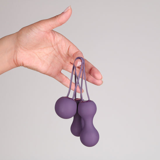 A hand holds the Je Joue Ami Kegel Ball Exerciser Set - Purple against a plain background. The set includes a silicone loop for easy handling, ideal for strengthening pelvic floor muscles.