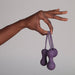 A hand gently holds the Je Joue Ami Kegel Ball Exerciser Set in purple, featuring three rounded silicone balls linked by loops. The plain gray background emphasizes their purpose in strengthening pelvic floor muscles.