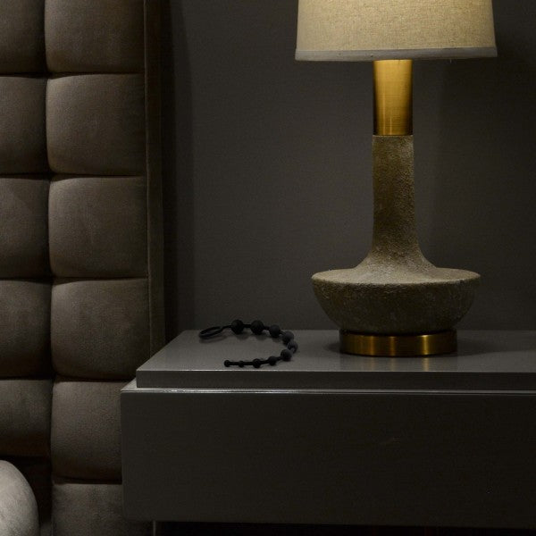 A modern bedside table featuring a textured grey lamp with a golden neck and cream-colored shade. Next to the lamp, there's a black beaded bracelet from Blush's Anal Adventures 10 Silicone Anal Beads, made from body-safe silicone material. The background displays a padded grey headboard. The scene is dimly lit, creating a cozy atmosphere.