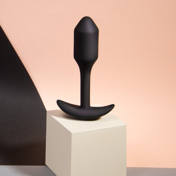 A B-vibe Snug Plug 1 Weighted Silicone Butt Plug - 2 oz - Black, featuring a curved base and slender neck, is displayed upright on a small beige pedestal. The background showcases soft, gradient colors transitioning from light peach to dark gray.