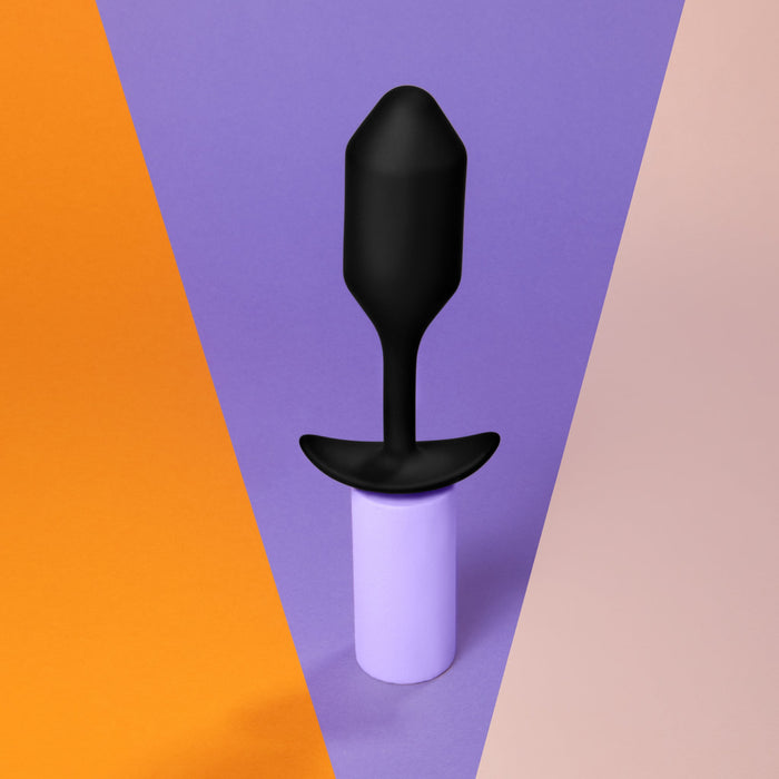 A b-Vibe Snug Plug 2 Weighted Silicone Butt Plug in black, featuring a flared base, is displayed upright and balanced on a purple cylinder. The background is divided into three colored sections: orange on the left, purple in the middle, and beige on the right.