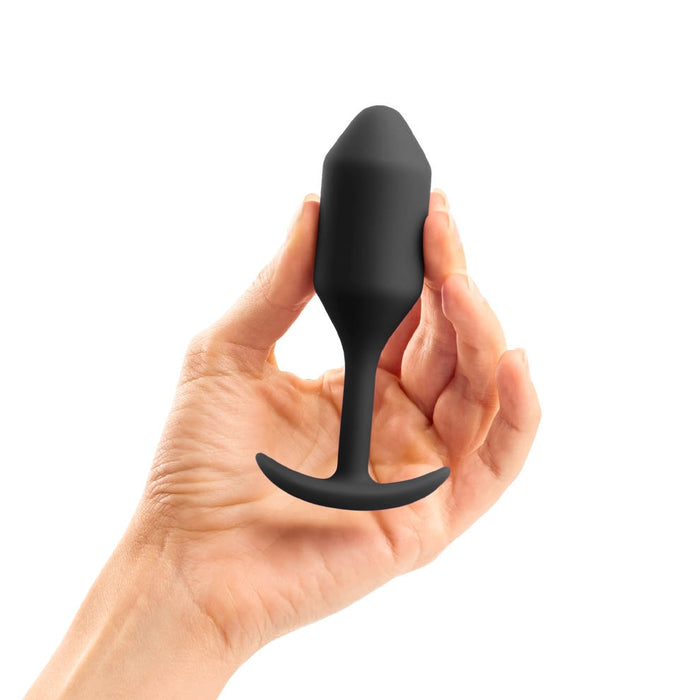 A hand holding the b-Vibe Snug Plug 2 Weighted Silicone Butt Plug - 5 oz - Black, with a tapered tip and flared base. This b-Vibe weighted plug is designed for safe and comfortable insertion. The background is plain white.