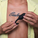 A person with a tattoo across their chest is holding a B-vibe Snug Plug 2 Weighted Silicone Butt Plug in black with both hands. The individual is wearing an open yellow shirt, exposing their tattooed chest underneath.