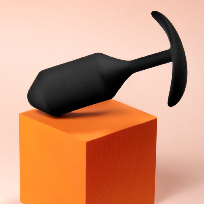 A sophisticated B-vibe Snug Plug 3 Weighted Silicone Butt Plug in black, featuring a T-shaped handle, is balanced diagonally on a vibrant orange cube against a soft peach background, creating an ideal visual for enhancing your anal play experience.