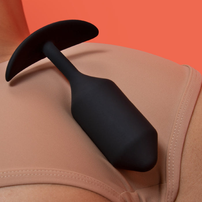 A B-vibe Snug Plug 3 Weighted Silicone Butt Plug - 6 oz, featuring a T-shaped handle, sits on the shoulder of a person wearing a beige top, set against a solid coral background.