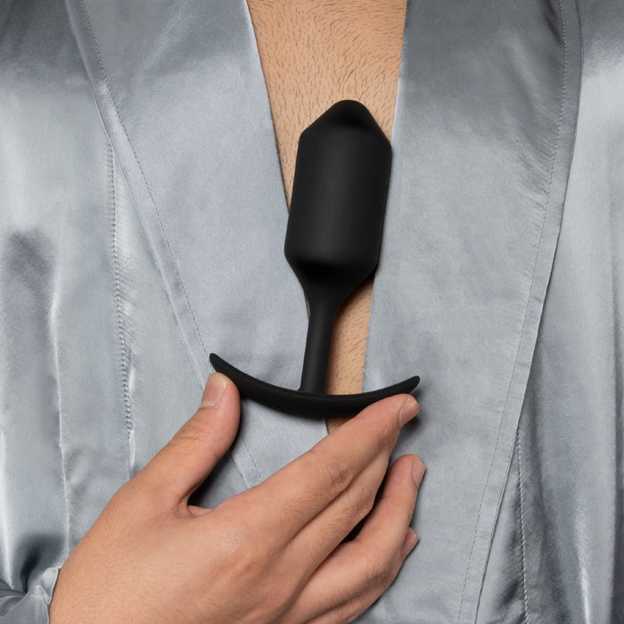 A person in a light gray satin robe holds the b-Vibe Snug Plug 4 XL Weighted Silicone Butt Plug (9 oz) - Black against their chest, with the focus on the person's hand and the plug.