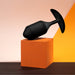 A sleek, black B-vibe Snug Plug 4 XL Weighted Silicone Butt Plug (9 oz) from b-Vibe rests on top of an orange geometric cube. The background features a gradient of soft pink and vibrant orange hues.