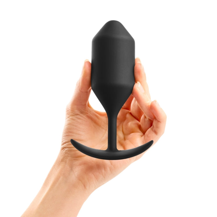 A hand holding a B-vibe Snug Plug 4 XL Weighted Silicone Butt Plug (9 oz), featuring a black silicone design with a tapered tip and curved base, ideal for weighted anal play, against a white background.