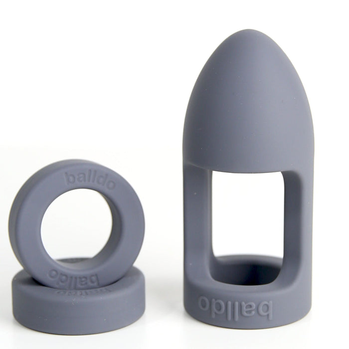 Gray silicone intimate accessories branded with "Balldo" on the side, featuring the Balldo: The World's First Ball Dildo - Grey. The set includes one open cylindrical ring and one larger, bullet-shaped piece with an open rectangular cutout, resembling a soft silicone cage. Both pieces are shown against a plain background, perfect for exploring ball orgasm sensations.