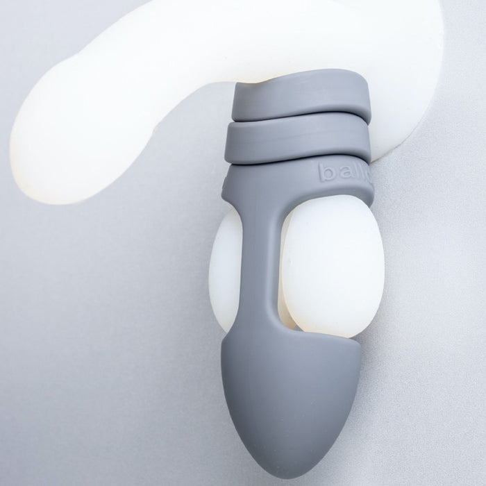 Close-up of the Balldo: The World's First Ball Dildo - Grey, featuring a snug, ergonomic design and soft contours, worn around a white, curved object. The product appears to have a comfortable fit, showcasing the innovative design by Balldo for intimate use. The background is a smooth, light gray surface.