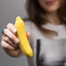 A person holds a small, yellow Banana Emojibator Ultra Discreet First Time Bullet Vibrator by Dame Products in their hand. The person's face is slightly blurred, featuring long brown hair and wearing a gray shirt. The silicone vibrator looks like a novelty item or accessory.