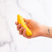 A hand with red and pink painted nails holds a small, yellow, silicone Banana Emojibator Ultra Discreet First Time Bullet Vibrator from Dame Products against a blurred white background. The wrist bears a tattoo with the word "love" written in cursive.