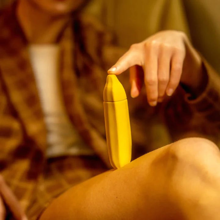 A person in a brown checkered outfit is holding a yellow, silicone, banana-shaped object—the Banana Emojibator Ultra Discreet First Time Bullet Vibrator by Dame Products—with their right hand, resting one finger on top of it. The object is placed on their crossed leg. The background is slightly blurred and in warm tones.