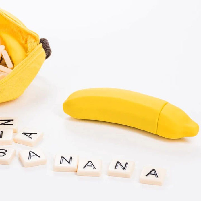 A yellow, silicone banana-shaped item, identified as the Banana Emojibator Ultra Discreet First Time Bullet Vibrator by Dame Products, is open with the zipper visible, showing wooden letter tiles inside. In front of it, tiles spell out "BANANA," placed in a row. The background is a clean white surface.