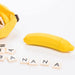 A yellow, silicone banana-shaped item, identified as the Banana Emojibator Ultra Discreet First Time Bullet Vibrator by Dame Products, is open with the zipper visible, showing wooden letter tiles inside. In front of it, tiles spell out "BANANA," placed in a row. The background is a clean white surface.