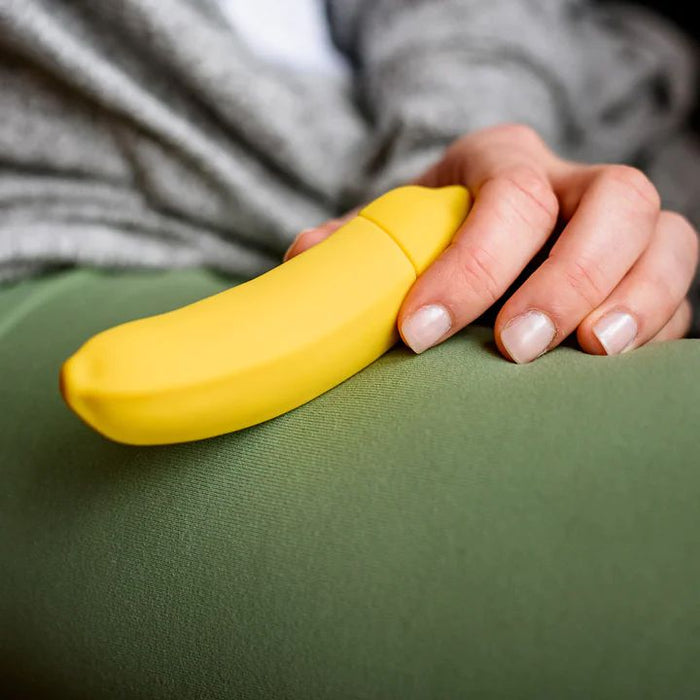 A person is holding a Banana Emojibator Ultra Discreet First Time Bullet Vibrator from Dame Products, made of silicone and shaped like a small, yellow banana. They are resting their hand on their leg, which is covered in green fabric, while wearing a gray garment. The object resembles a novelty item or toy and might be a vibrating toy.