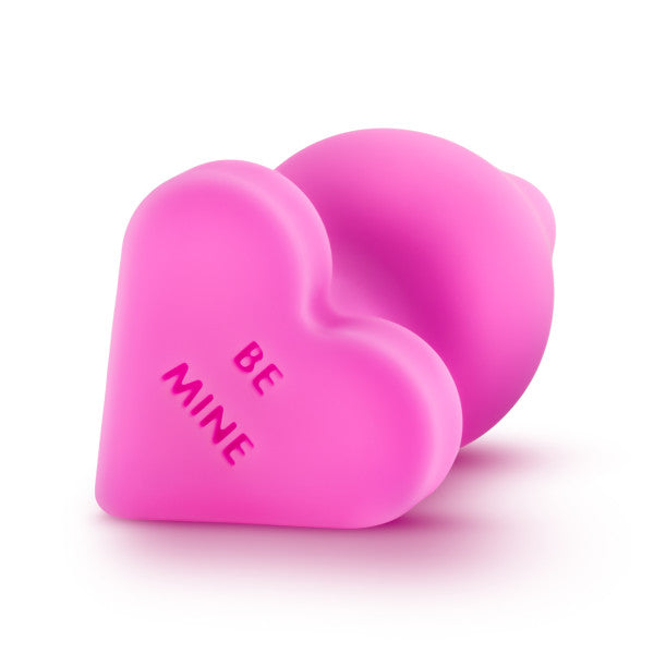 An image of two bright pink Be Mine Naughty Candy Heart Butt Plugs by Blush, with one standing upright and the other lying on its side. The upright heart is embossed with the words "BE MINE". Crafted from soft silicone, their surfaces are smooth and cast shadows beneath them against a white background.