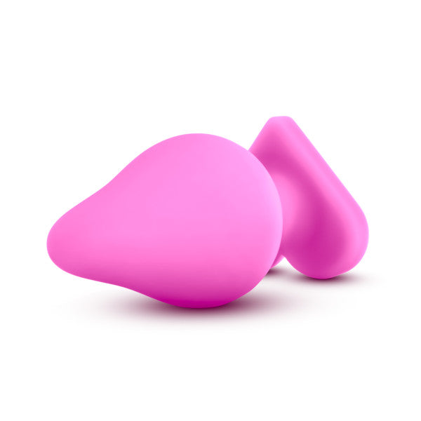 A pair of Be Mine Naughty Candy Heart Butt Plugs by Blush, crafted from soft pink silicone and featuring a glossy, smooth finish, are placed against a white background. One plug is slightly larger and rounder in shape, while the other has a more triangular form. These heart-shaped plugs have an abstract design that makes them perfect for anal beginners.