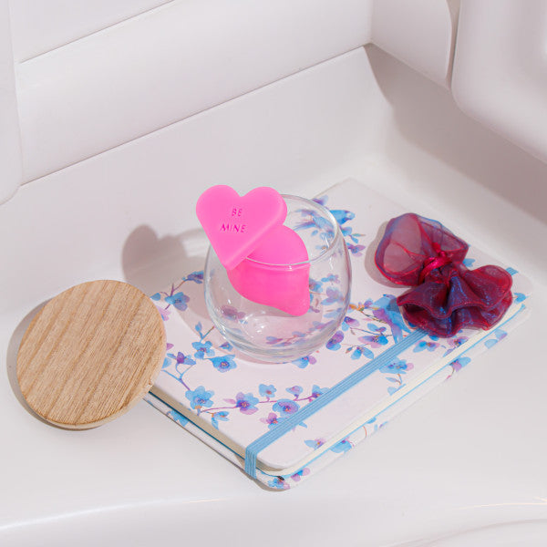 A desk featuring a floral-patterned notebook, a wooden coaster, a small round glass container with a pink heart-shaped tag that reads "BE MINE," and two hair scrunchies, one red and one bluish-purple. Off to the side is the Be Mine Naughty Candy Heart Butt Plug in Pink by Blush, designed with soft silicone for anal beginners.