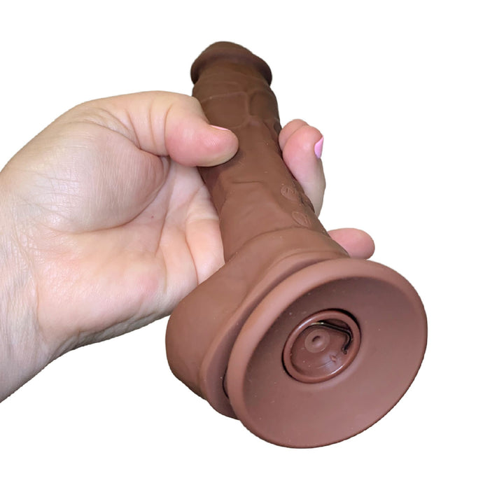 A person's hand is gripping a brown cylindrical object with a flared base, closely resembling the Big Shot Blaster 8 Inch Vibrating Squirting Silicone Dildo (Ejaculating Strap on) in Chocolate by Betty's Toy Box. The background is white, highlighting the grip and part of the object.