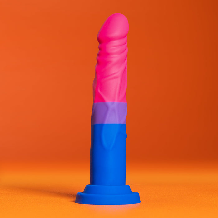 The Bisexual Pride 7.5 Inch Silicone Suction Cup Dildo from Blush stands upright against an orange background. This multicolored, phallic-shaped object features a blue base, purple midsection, and pink tip, crafted from platinum cured silicone to proudly display the hues of the bisexual flag.