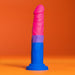 The Bisexual Pride 7.5 Inch Silicone Suction Cup Dildo from Blush stands upright against an orange background. This multicolored, phallic-shaped object features a blue base, purple midsection, and pink tip, crafted from platinum cured silicone to proudly display the hues of the bisexual flag.