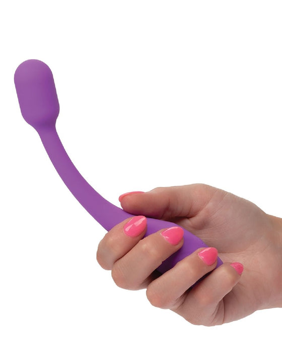 A hand with pink-painted nails holds the Bliss Flex O Teaser Slim First Time G Spot Vibrator by CalExotics, a purple, curved wand-shaped object with a round bulbous end, perfect for G-spot exploration.