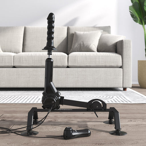 The Body Dock Thrusting Sex Machine with Remote from Pipedream Products, featuring a sleek black design and equipped with cables and a compact control unit, is positioned on a wooden floor in the living room. A gray sofa and a large plant enhance the backdrop as the device stands ready for action.