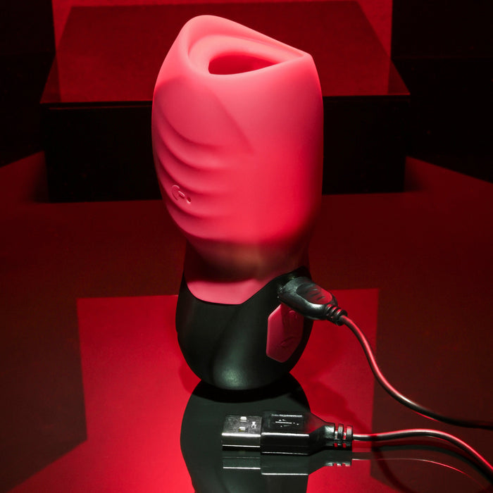 The Evolved Novelties Body Kisses Red Suction Masturbator and Vibrator, featuring a red and black design, textured grips, an opening at the top, and USB cable plugged in, is placed on a reflective red surface against a dark background. This cylindrical electronic device offers 7 speeds for customized use.