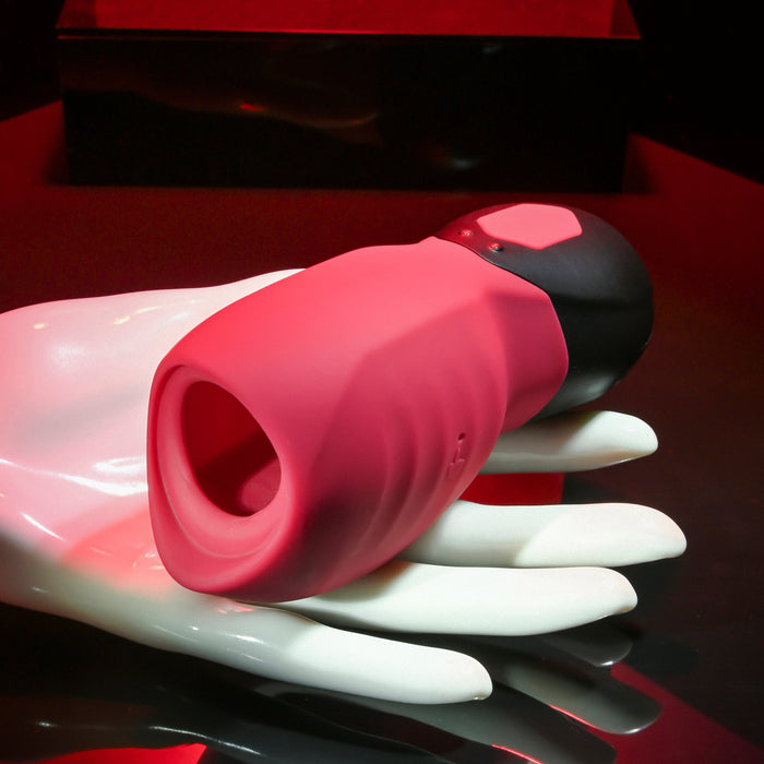 A white mannequin hand holds the Body Kisses Red Suction Masturbator and Vibrator by Evolved Novelties, showcasing its pink and black design with a rounded shape and hollow opening. The creamy silicone device, which features a vibrating reverse side, is set against a dark background illuminated with red highlights, suggesting a sleek and modern design.