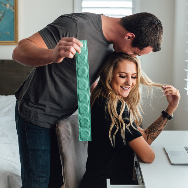 A man kisses a woman's head while she smiles at her desk in what seems to be a bedroom, holding a long green package of Royal Condom Extra Large Vegan Condoms 20-Pack.