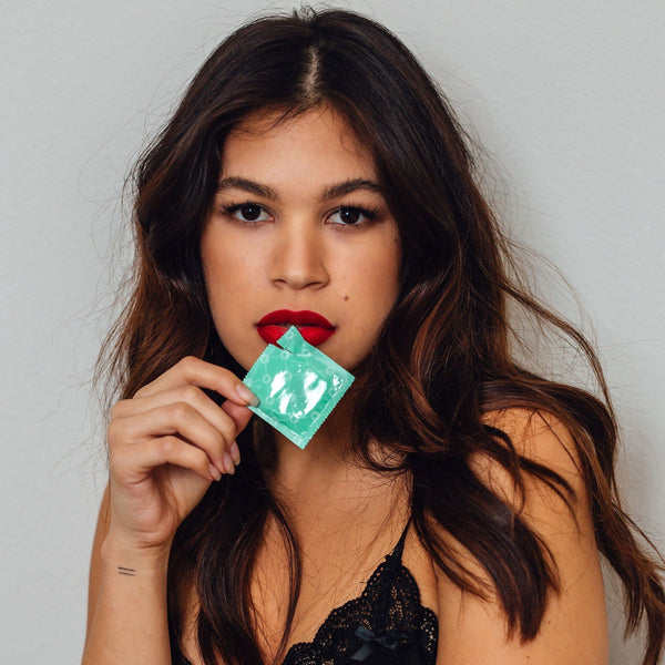 A person with long brown hair and red lipstick holds a green packet of Royal Condom Extra Large Vegan Condoms 20-Pack against a plain background, wearing a black top and looking directly at the camera.