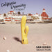 A yellow California Dreaming San Diego Seduction G-Spot Thumping Rabbit Vibrator by CalExotics stands upright on a sandy beach near seashells. In the background, there is a large historic hotel with red roof tiles, surrounded by palm trees under blue skies. The image text reads "California Dreaming by CalExotics" and "San Diego Seduction.