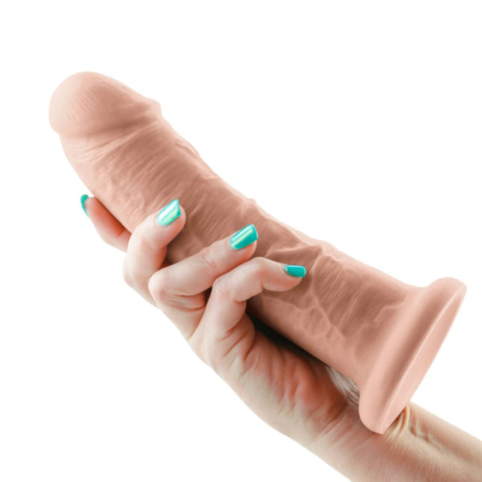 A hand with teal-colored nails holds an NS Novelties Colours Girthy 8 Inch Dual Density Silicone Suction Cup Dildo - Vanilla against a white background. The dildo has a textured surface and distinct head and veins, giving it an anatomically accurate appearance.