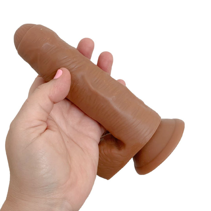 A hand holds the Coverboy Derek the Bartender 7 Inch Uncut Dildo in Mocha by Blush, featuring a phallic shape crafted from a soft, body-safe material with a realistic skin texture and a suction cup base. The background is plain white.