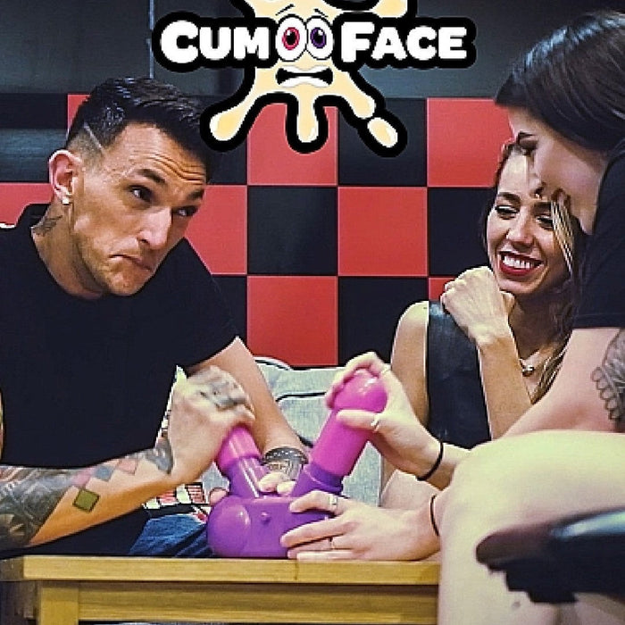 Three people are seated around a table playing a game. The man on the left and the woman on the right are holding and aiming pink water guns at a purple device. A smiling woman sits in the middle. Above them is a logo with the text "Cum Face Duel Pump Action Penis Game" and a splash graphic with eyes—an ideal party piece for any bachelor party by Shots.