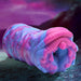 A close-up photograph of a fantasy-themed, colorful silicone toy. The Cyclone Squishy Alien Vagina Role Play Stroker by XR Brands is predominantly blue and pink with a textured, ridged, and ruffled surface, resembling an abstract, organic form. The background is a mystical scene with purple and blue hues.