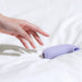 A hand reaching towards a small, smooth Dame Aer Clitoral Pressure Wave Vibrator - Purple from Dame Products on a white surface. The device, designed for clitoral stimulation, boasts an ergonomic design and a simple, sleek appearance.