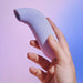 A hand holds a Dame Aer Clitoral Pressure Wave Vibrator - Purple from Dame Products against a background with a gradient from pink to blue. Renowned for its pressure wave technology, this ergonomically-shaped device has a rounded, slightly curved body with a circular opening at one end. The scene is softly lit, giving the vibrator a smooth appearance.