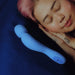 A person with pink hair is sleeping peacefully with their hands under their cheek. Beside them, on a blue pillow, is a Dame Com Ergonomic Waterproof Wand Vibrator in Periwinkle by Dame Products. The overall atmosphere is calm and soothing.