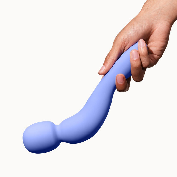 A hand is holding the Dame Com Ergonomic Waterproof Wand Vibrator by Dame Products, a periwinkle curved silicone device designed for clitoral stimulation with a rounded, bulbous end against a white background.