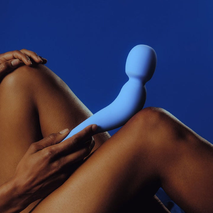 A close-up of a person holding the Dame Com Ergonomic Waterproof Wand Vibrator in Periwinkle against their bare legs, set against a deep blue background. The body-friendly wand, designed by Dame Products, appears to be a personal care device. The person's hand gently grips the massager made of medical grade silicone, which contrasts with their skin tone.