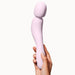 A hand holding the Dame Com Ergonomic Waterproof Wand Vibrator - Quartz by Dame Products against a plain white background. The light pink, curved wand-shaped device appears smooth and ergonomically designed with a rounded tip and mid-length curve, made from medical grade silicone for safe clitoral stimulation.