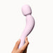 A hand holding the Dame Com Ergonomic Waterproof Wand Vibrator in Quartz, a light pink personal massager by Dame Products, made of medical grade silicone against a white background. The device, ideal for clitoral stimulation, features a rounded head and a smooth surface.