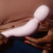 A person with red-painted nails holds the Dame Com Ergonomic Waterproof Wand Vibrator - Quartz by Dame Products, an abstract soft pink sculpture made of medical grade silicone, with both hands. The background shows part of the person's torso and part of a garment, creating a warm color contrast.