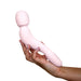 A hand is holding the Dame Com Ergonomic Waterproof Wand Vibrator in Quartz, a flesh-colored, L-shaped personal massager from Dame Products. It features a rounded, bulbous head and a curved handle made from medical grade silicone. The background is white.