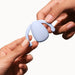 Two hands are shown holding the Dame Eva Hands-Free Silicone Clitoral Vibrator - Ice Blue by Dame Products against a plain white background. The object appears to be a small, smooth device with a teardrop shape and flexible arms. One hand holds the device from the bottom, while the other grips it from the top.