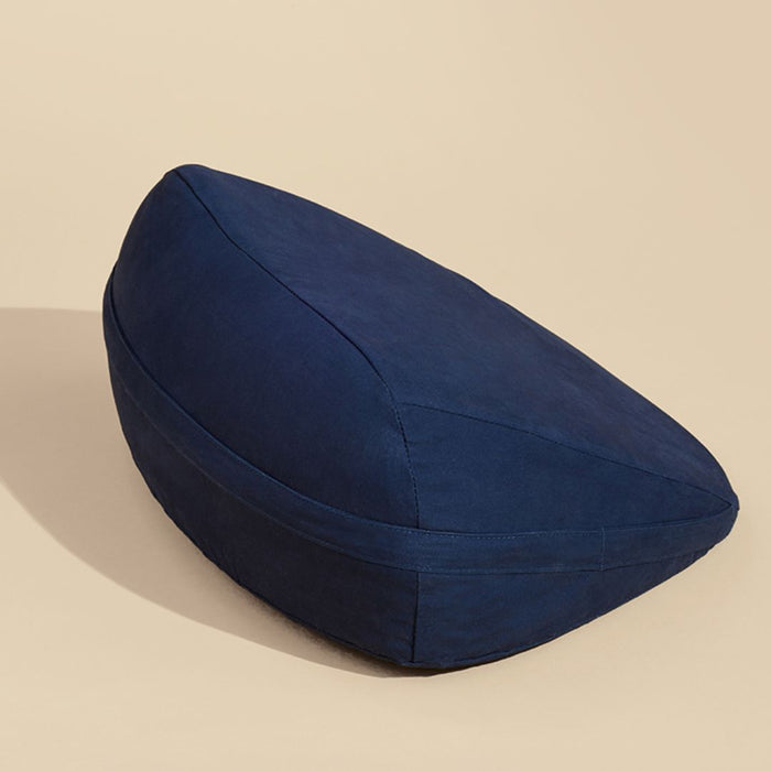 A dark blue, wedge-shaped Dame Pillo Sex Pillow for Sex Positioning - Indigo by Dame Products is placed against a beige background. The cushion appears to be made of soft fabric and is slightly elevated on one side, casting a small shadow.