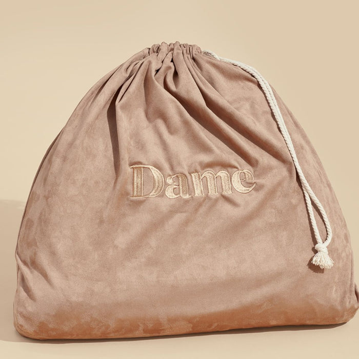 A soft, pinkish-brown drawstring bag with the word "Dame Products" embroidered on the front. The velvety material and white rope for the drawstring give it an elegant touch. Perfect for discreetly storing your intimate accessories, such as the Dame Pillo Sex Pillow for Sex Positioning - Indigo, against a beige background.